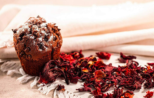 Ein saftiger Schokoladenmuffin neben getrockneten Hibiskusblüten auf einer rustikalen Unterlage, einladend zum Genuss