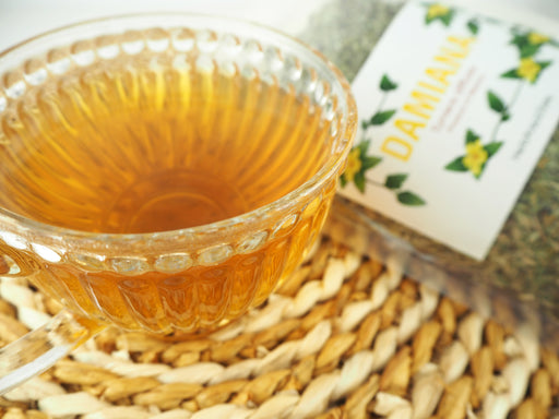 Eine Tasse goldener Damianatee neben einer Verpackung des Tees, betont durch die gelben Blüten des Turnera diffusa Strauchs