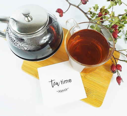 Teezeit-Setup mit einer klaren Teekanne, Tasse mit heißem Tee und einer Karte mit Aufschrift 'Tea time', umgeben von Hagebuttenzweigen.