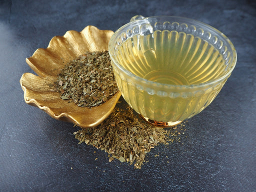 Goldene Schale mit getrockneten Ginkgo Biloba Blättern neben einer klaren Teetasse, auf dunklem Untergrund.