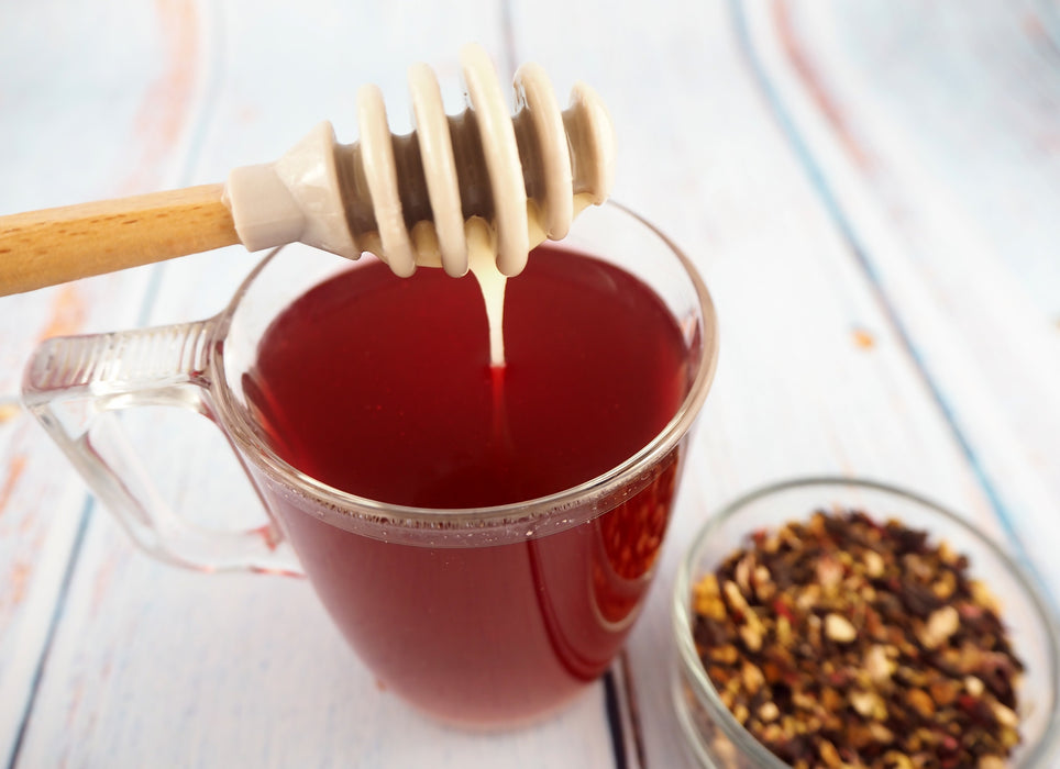 Honig tropft von einem Holzlöffel in eine Tasse mit rotem Kräutertee, begleitet von einer entspannenden Kerze im Hintergrund.