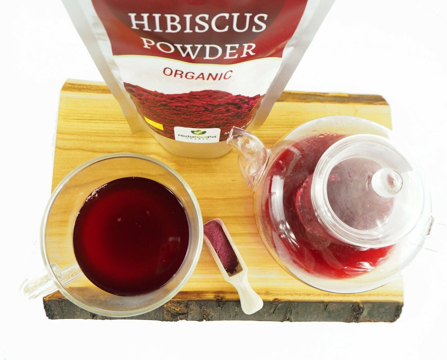 Organisches Hibiskuspulver in einer transparenten Verpackung auf einem Holzbrett, daneben eine gefüllte Tasse roten Tees und eine gläserne Teekanne
