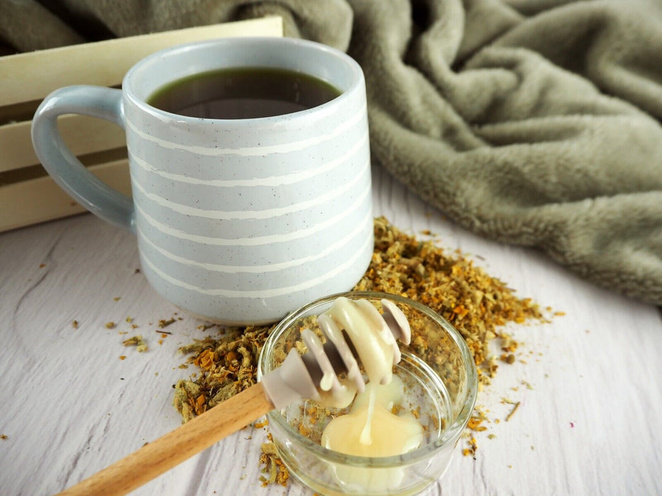 Eine große, gemusterte Tasse mit heißem Tee neben einer Schale mit losen Kräutern und einem Honiglöffel auf einem Holzbrett.