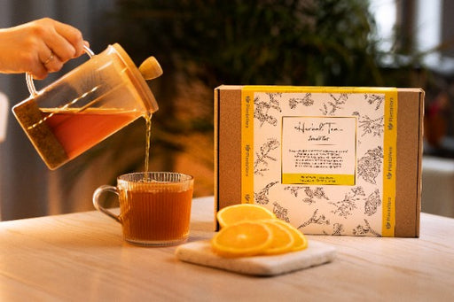 Teekanne gießt frisch aufgebrühten Kräutertee in eine Tasse neben einer Geschenkbox mit Orangenscheibe auf hellem Holztisch.