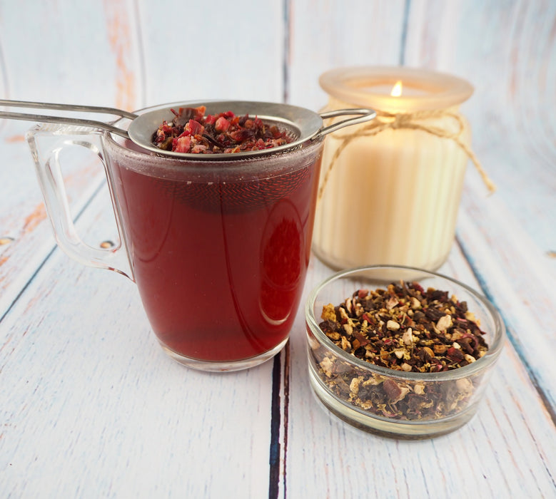 Transparente Teetasse mit rotem Kräutertee, daneben eine Schale mit losen Teeblättern und eine brennende Kerze.