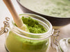Grünes Bio Moringa Pulver in einem Glasgefäß mit Schließverschluss und einem Stößel auf einem hellen Küchentisch.