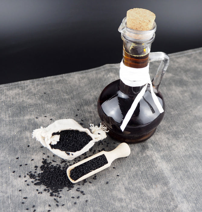 Ein dunkles Fläschchen mit Bio Schwarzkümmelöl neben Schwarzkümmelsamen auf einem grauen Tuch, arrangiert für ein natürliches Produktbild.