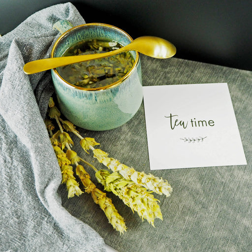 Bild einer Tasse Bergtee auf einem grauen Leinentuch, neben einer Karte mit der Aufschrift „Tea time“ und einem Bündel getrockneter Blumen