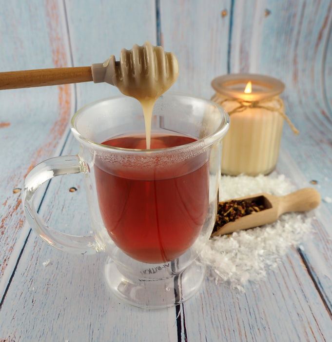 Glasklarer Becher gefüllt mit rotem Kräutertee und Honiglöffel darüber, auf einem Holztisch mit winterlichem Dekor.